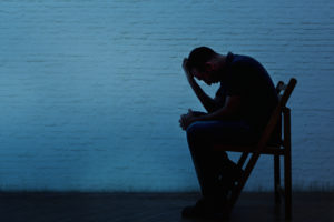 Unhappy and lonely young man in front of a wall. He sits on a chair and thinks alone under blue light. Negative human expression.